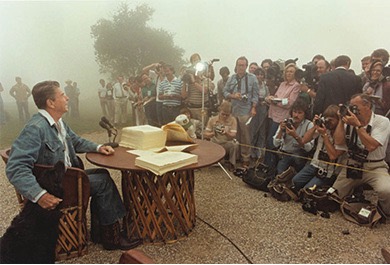 A color photo of Ronald Reagan seated at a table outdoors with papers on it at his ranch. He is wearing blue jeans, denim jacket, and cowboy boots and pets his dog who is beside his chair.  The press corp and others face him.