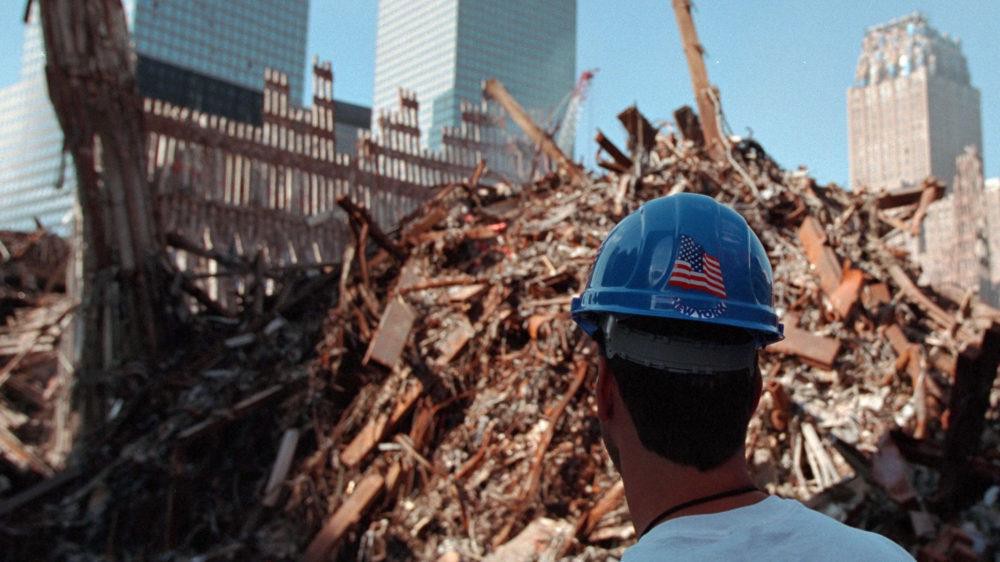 Photo of a worker wearing a hard hat with the American Flag emblem on the back looks at the rubble of and remains of the World Trade Center