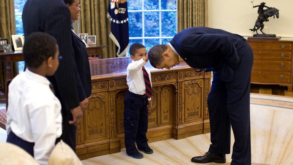 Photo of President Barak Obama and young boy standing in front of the President's desk in the Oval office. The president is bending down so the young boy can touch his hair.
