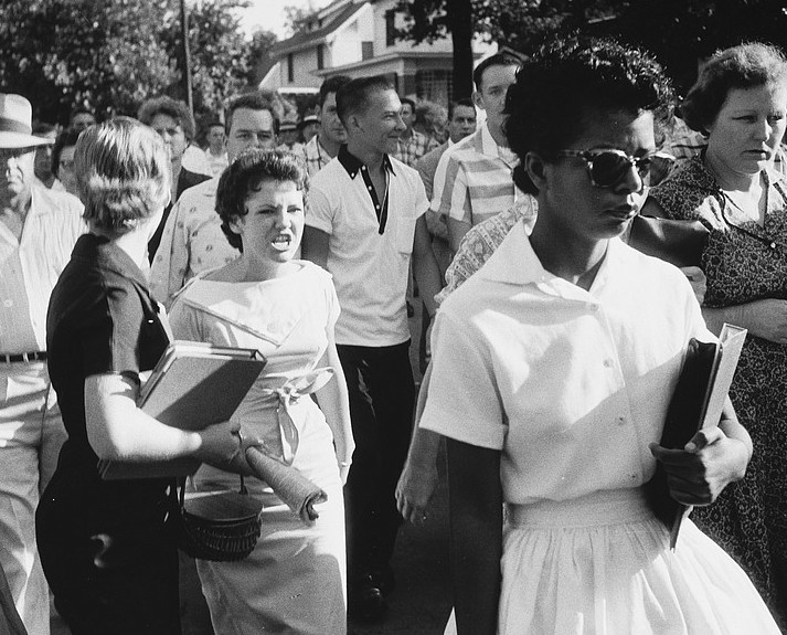 black and white photograph of Elizabeth Eckford wearing a white blouse and skirt, wearing sunglasses, and carrying a textbook while being surrounded by an angry crowd of white people.